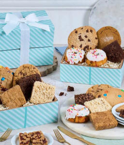 Two rainbow striped gift boxes with a blue bow. In front, a plate with a mini bundt cake with a candle, another tray with cookies, brownies, and blondies. Behind those, a cake tray with whoopie pies and mini bundt cakes. 